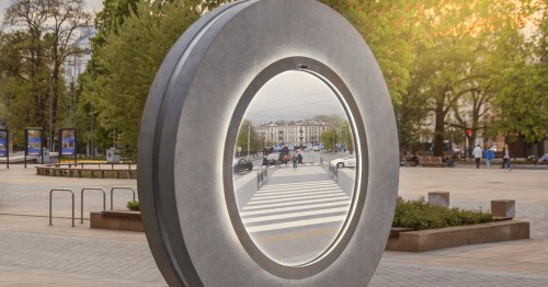 A large circular mirror sculpture reflects a city street with pedestrians and greenery in the background.