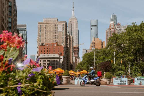 A city street scene with flowers in the foreground, featuring the Empire State Building and outdoor dining in the background.