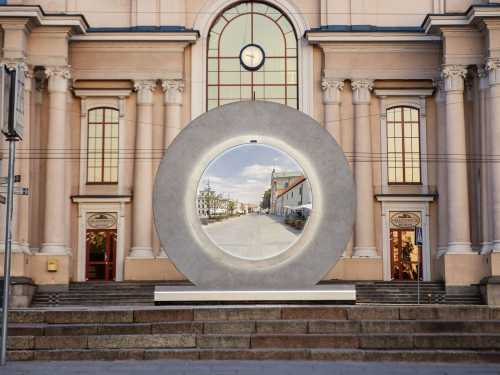 A large circular frame stands in front of a building, showcasing a view of a street scene through it.