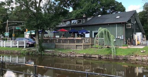 A green building with a deck and umbrellas by a calm river, surrounded by trees and a road in the background.