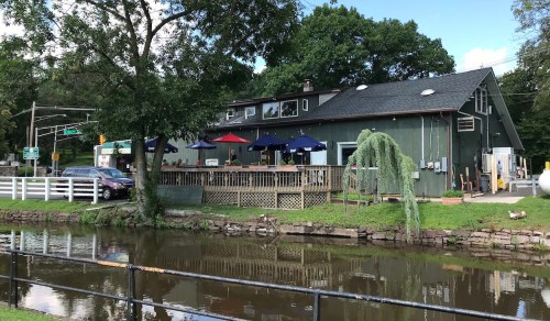 A green building with a deck and umbrellas, beside a calm waterway, surrounded by trees and a road in the background.