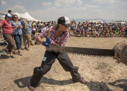 A woman in a plaid shirt and cap saws a log in front of a cheering crowd at an outdoor event.