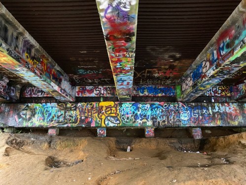 Colorful graffiti covers the pillars and underside of a bridge, with sandy ground visible below.