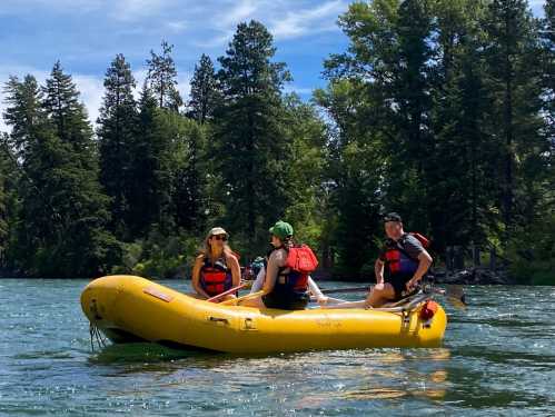 Three people in a yellow raft on a river, surrounded by trees and a clear blue sky.