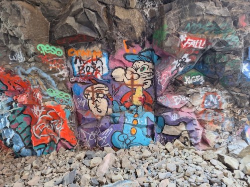 Colorful graffiti on a rocky wall, featuring cartoon characters and abstract designs amidst scattered stones.