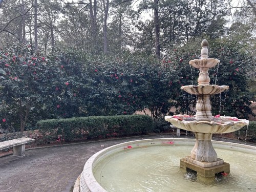A stone fountain with three tiers, surrounded by lush greenery and blooming flowers in a serene garden setting.