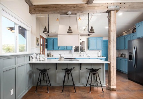 Modern kitchen with blue cabinets, a white countertop, and bar stools, featuring wooden beams and pendant lighting.