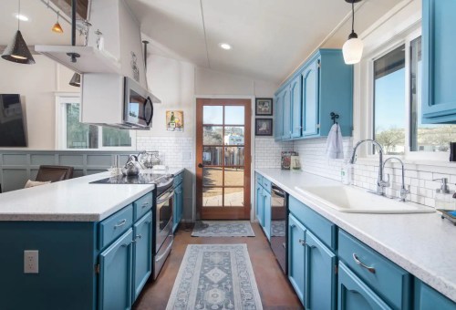Bright kitchen with blue cabinets, white countertops, and a door leading to an outdoor area, featuring natural light.