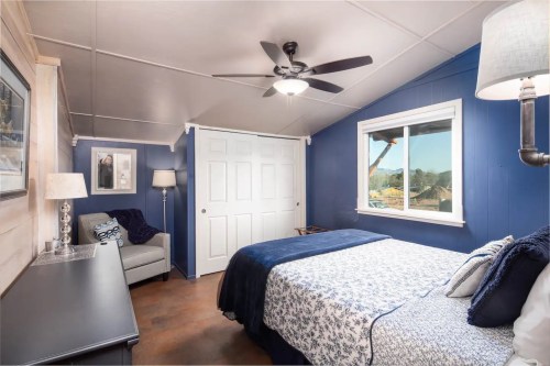 Cozy bedroom with blue walls, a queen bed, a chair, and a ceiling fan, featuring natural light from a window.