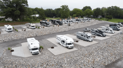Aerial view of a parking area with multiple RVs and campers arranged on concrete pads surrounded by gravel and greenery.
