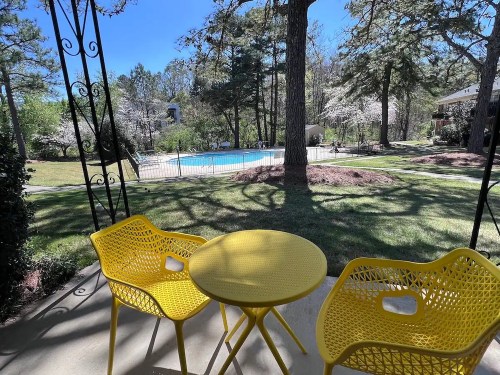 A sunny outdoor view featuring a yellow table and chairs overlooking a pool surrounded by trees and greenery.