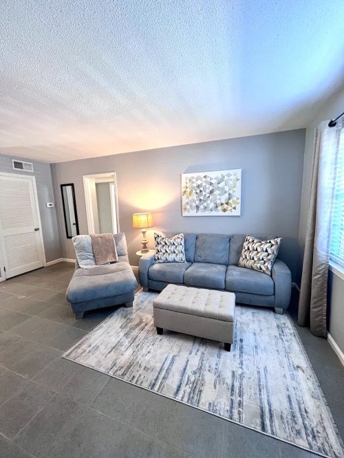 Cozy living room with a blue sofa, armchair, ottoman, and decorative rug against a light gray wall.