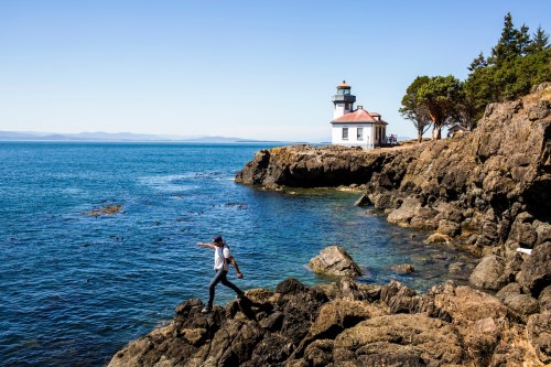 A person balances on rocky shore near a lighthouse, with calm blue waters and clear skies in the background.