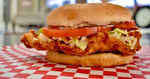 A crispy fried chicken sandwich with lettuce and tomato on a toasted bun, served on a red and white checkered paper.