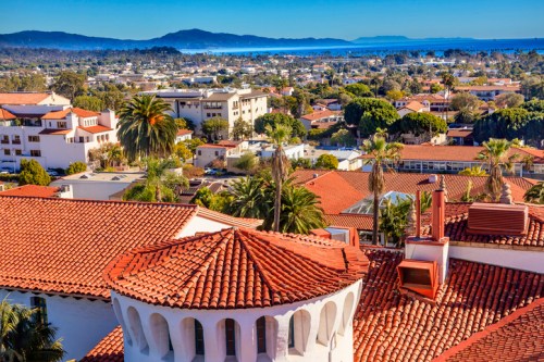 A scenic view of a coastal town with red-tiled roofs, palm trees, and mountains in the background under a clear blue sky.