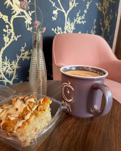 A cozy scene featuring a slice of cake, a coffee mug, and a vase with flowers on a wooden table beside a pink chair.