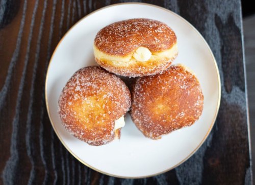 Three sugar-coated donuts filled with cream, arranged on a white plate atop a dark wooden surface.