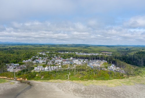 Aerial view of a coastal town surrounded by green forests and sandy beach under a cloudy sky.
