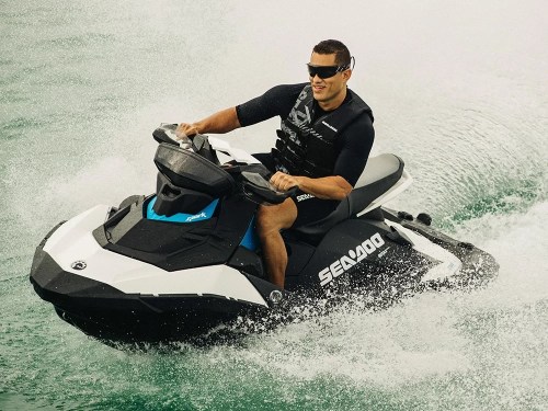 A person riding a Sea-Doo jet ski on water, wearing sunglasses and a life jacket, with splashes around.