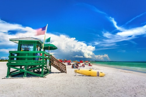 A vibrant beach scene featuring a green lifeguard tower, sandy shore, and colorful umbrellas under a blue sky.