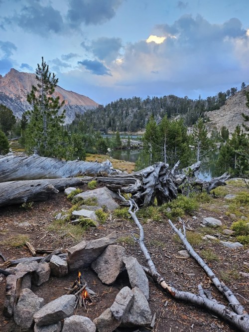 A serene mountain landscape with a small campfire, fallen logs, and a lake surrounded by trees under a cloudy sky.