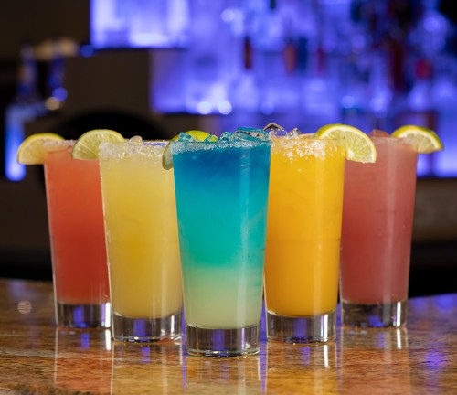 Five colorful cocktails with garnished lime slices, arranged on a bar counter with a vibrant background.