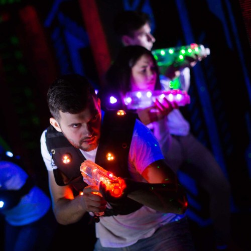 A group of people in dark attire play laser tag, holding illuminated laser guns in a colorful, neon-lit environment.