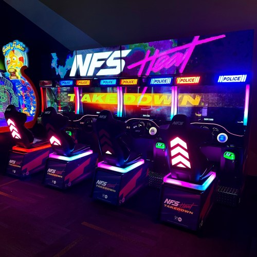 A row of colorful arcade racing games titled "NFS Heat: Takedown" with illuminated screens and seats.