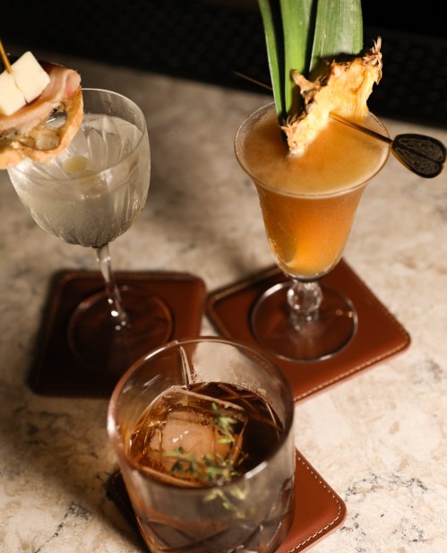 Three stylish cocktails on a marble surface, featuring a fruity drink with a pineapple garnish and two other elegant glasses.