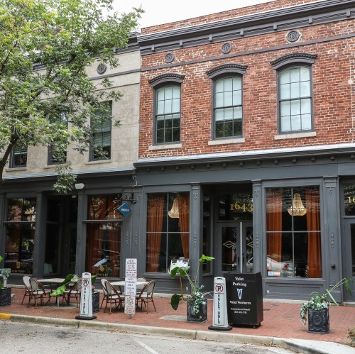 Historic brick building with large windows, outdoor seating, and decorative elements, surrounded by trees.