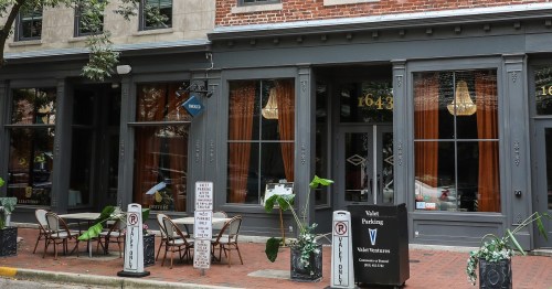 Exterior of a restaurant with large windows, outdoor seating, and potted plants, featuring a brick facade and elegant decor.