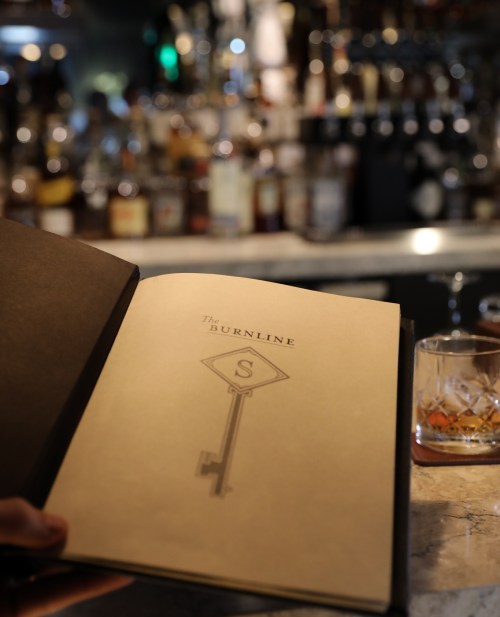 A hand holds an open menu titled "The Burnline" in a dimly lit bar with bottles in the background.