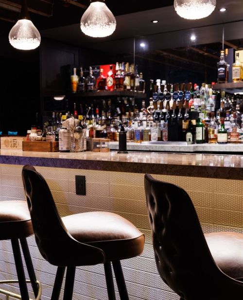 A stylish bar with three leather stools and a well-stocked shelf of various liquor bottles in the background.