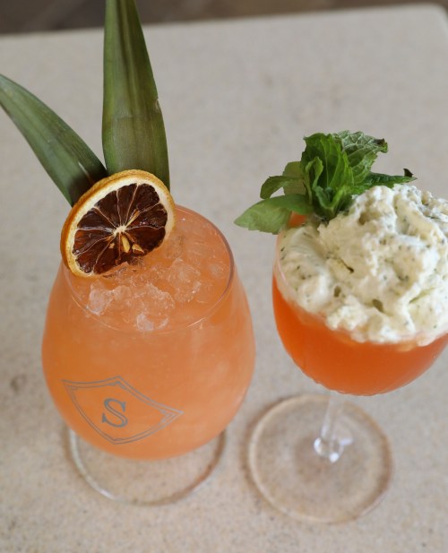 Two colorful cocktails on a light surface: one with pineapple and dried citrus, the other topped with mint and cream.