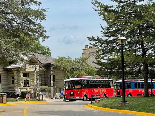 A red trolley bus parked near a log cabin-style building surrounded by trees on a sunny day.