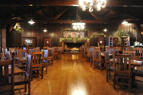 A rustic dining room with wooden tables and chairs, a large fireplace, and decorative greenery on the walls.