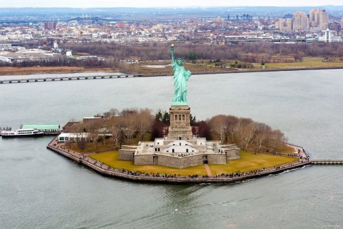 Aerial view of the Statue of Liberty on Liberty Island, surrounded by water and city skyline in the background.