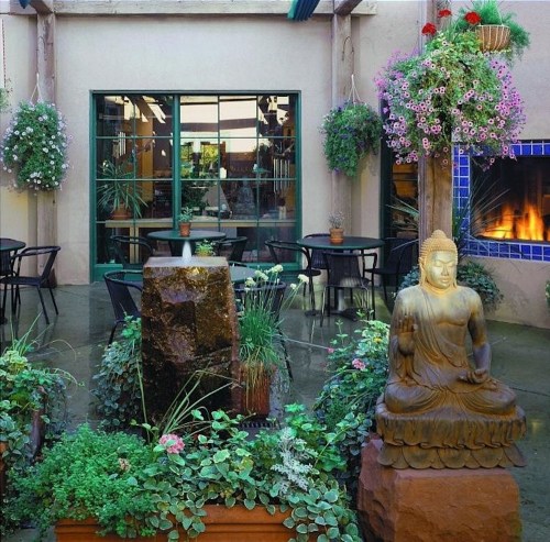 A serene courtyard with a Buddha statue, lush greenery, potted plants, and a cozy fireplace in the background.