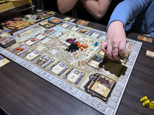 A tabletop game in progress, featuring a detailed board, various tokens, and a player's hand placing a card.