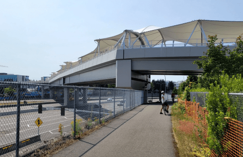A pedestrian path alongside a highway, with a modern elevated structure covered by white canopies above.