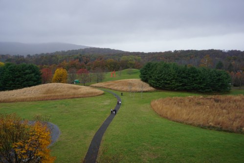 A winding path through a grassy landscape with trees and rolling hills, under a cloudy sky. Autumn colors are visible.