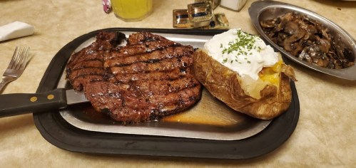A grilled steak on a platter with a baked potato topped with sour cream and chives, alongside a dish of mushrooms.