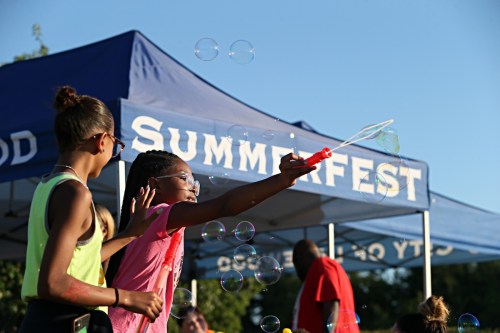 Two children play with bubbles in front of a blue tent labeled "Summerfest" on a sunny day.