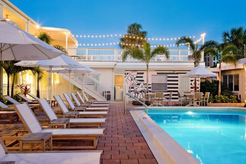A serene pool area with lounge chairs, umbrellas, and string lights, surrounded by palm trees at dusk.