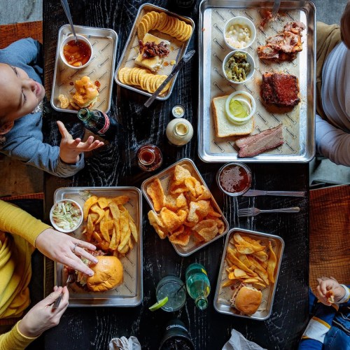 Aerial view of a table with various BBQ dishes, sides, and drinks, with hands reaching for food.