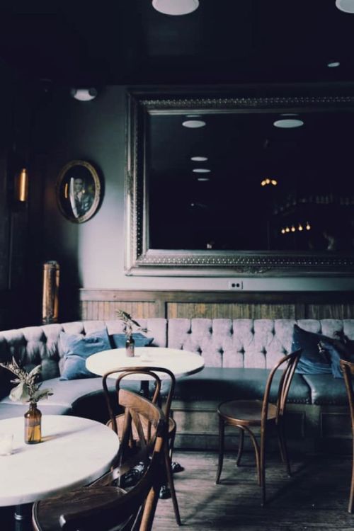 Cozy café interior with a tufted gray sofa, wooden chairs, and a large mirror reflecting soft lighting.
