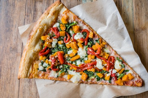 A large slice of pizza topped with spinach, red peppers, butternut squash, and cheese on a brown paper background.