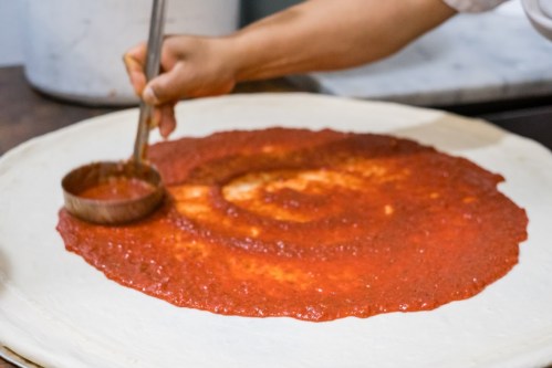 A hand spreads tomato sauce in a spiral pattern on a large pizza dough.
