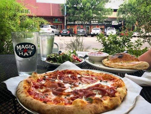 A table outdoors featuring a pizza with ham, a salad, and drinks, with shops and greenery in the background.
