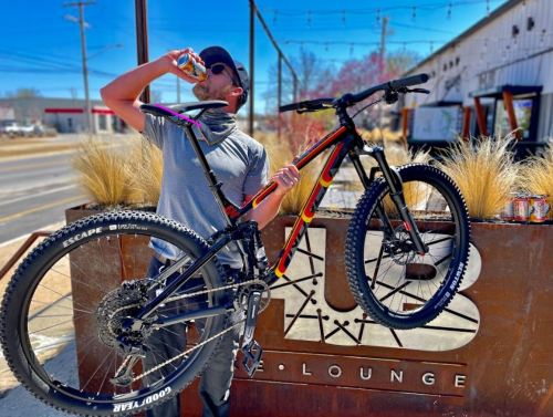 A person drinks from a can while holding a mountain bike in front of a lounge sign on a sunny day.